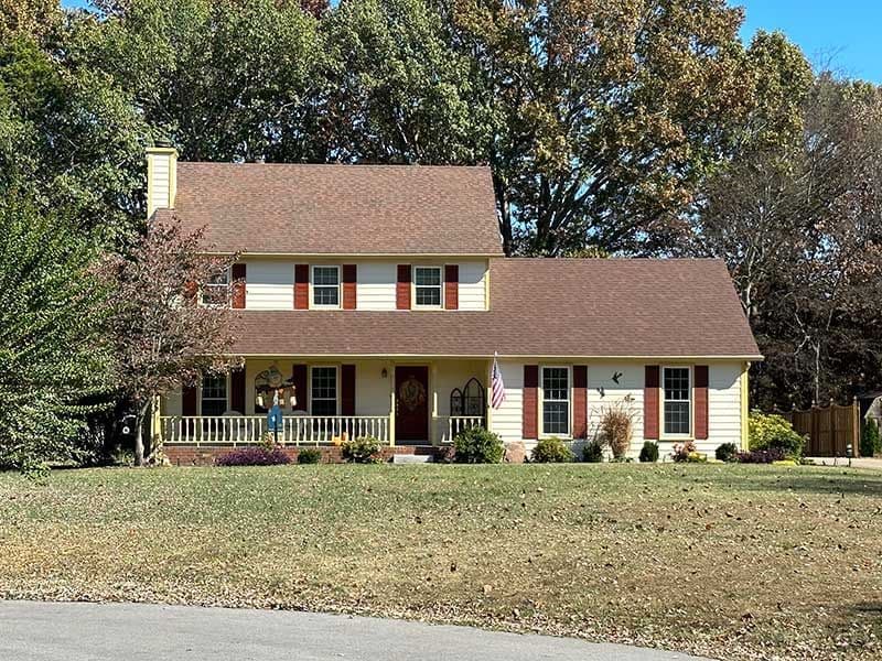 Two-story yellow house with brown roof, surrounded by trees and autumn foliage.