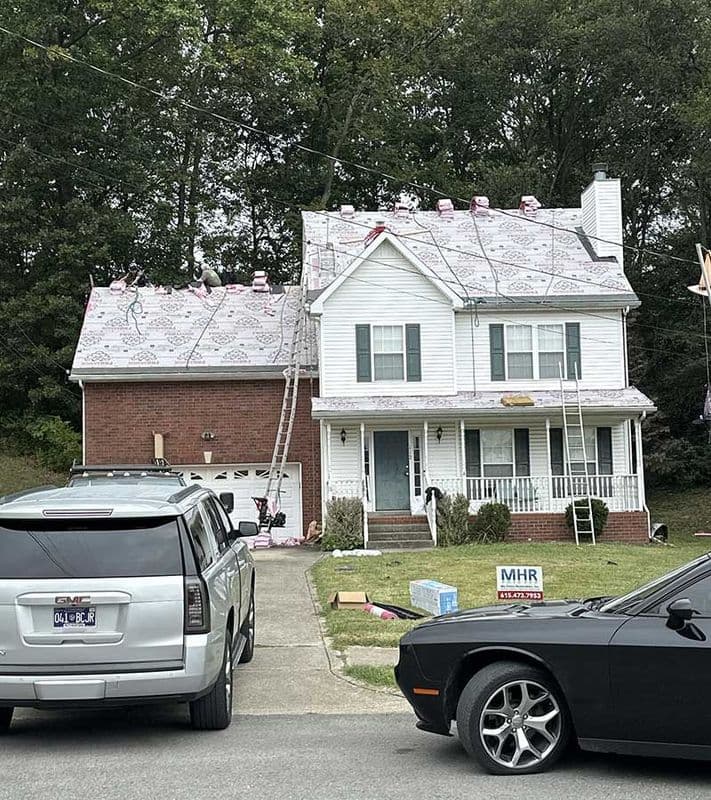 Roofing installation on a two-story house with construction workers and equipment visible.